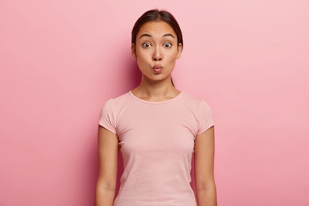 Mooie Aziatische meid met een gezonde huid, natuurlijke schoonheid, houdt de lippen gevouwen voor een kus, trekt wenkbrauwen op, heeft donker haar dat in een paardenstaart is gekamd, draagt een casual outfit, modellen over een roze muur. Menselijke gezichtsuitdrukking