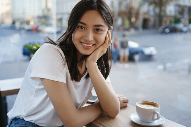 Mooie Aziatische meisje koffietafel zitten, cappucino drinken en glimlachen.