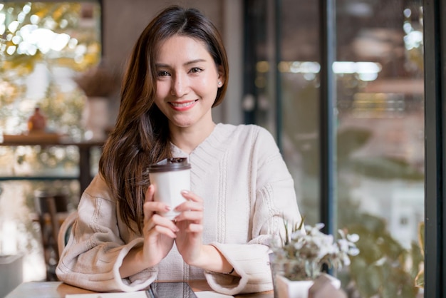 Mooie aziatische vrouw geniet van een warme drank in de buurt van het raam in het concept van de levensstijlideeën van de caféwinkel