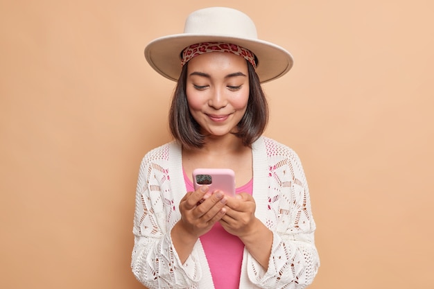 Mooie aziatische vrouw met donker haar houdt moderne mobiele telefoon controleert bericht verbonden met draadloos internet draagt fedora gebreide witte jas geïsoleerd over beige muur maakt gebruik van mobiele applicatie