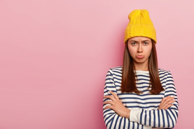 Mooie beledigde vrouw kijkt met een bedroefde uitdrukking, fronst haar wenkbrauwen en tuit de lippen van ongenoegen, houdt armen over elkaar, draagt stijlvolle gele hoed en gestreepte trui, modellen over roze muur, lege ruimte