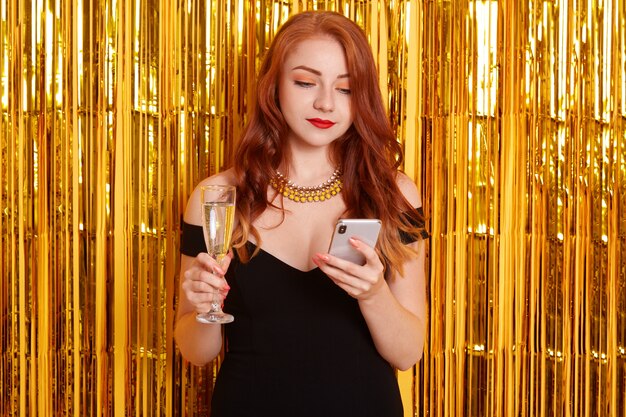 Mooie blanke vrouw chats op telefoon en wijn drinken, ziet er geconcentreerd, roodharige dame met krulspelden permanent geïsoleerd over gouden klatergoud, vrouw met slimme telefoon.