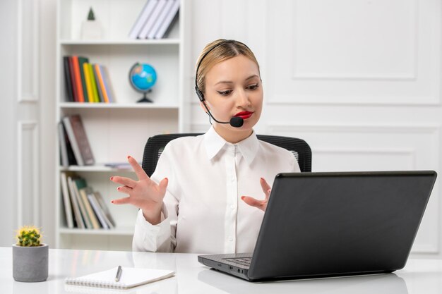 Mooie blonde meid van de klantenservice in een wit overhemd met een laptop en een headset die met de handen zwaait en luistert