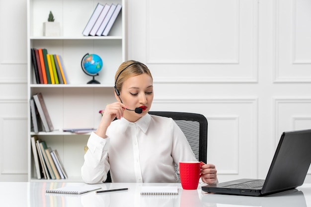 Mooie blonde meid van de klantenservice in een wit overhemd met een laptop en een headset die op een microfoon praat