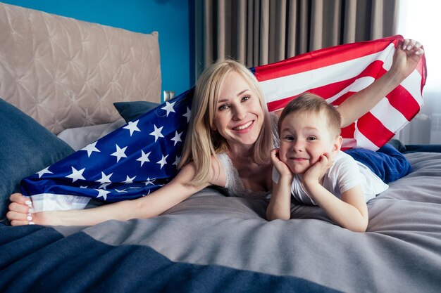 Mooie blonde moeder en zoon liggend op het bed in de slaapkamer met amerikaanse vlag in een comfortabel appartement safe house.4 juli onafhankelijkheidsdag viering concept en engels leren