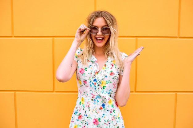 Mooie blonde vrouw die bloemenkleding draagt, die dichtbij gele muur stelt