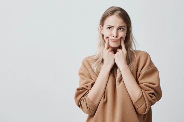 mooie blonde vrouw die grimas maakt, haar wangen met vingers aanraakt, naar boven kijkend, met een ontevreden uitdrukking. Gezichtsuitdrukking en negatieve emoties