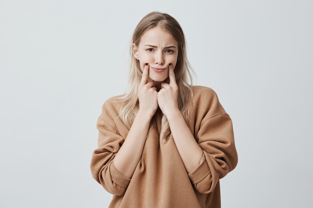 mooie blonde vrouw die grimas maakt, haar wangen met vingers aanraakt, zichzelf laat glimlachen, fronsend gezicht, ontstemd en overstuur. Gezichtsuitdrukking en negatieve emoties