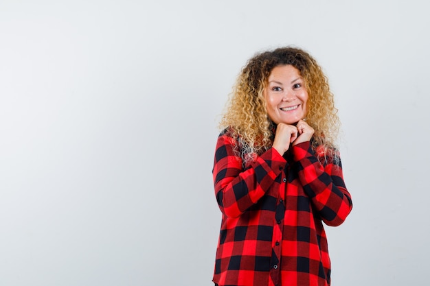 Mooie blonde vrouw die haar handen onder de kin houdt in een geruit overhemd en er beschaamd uitziet. vooraanzicht.