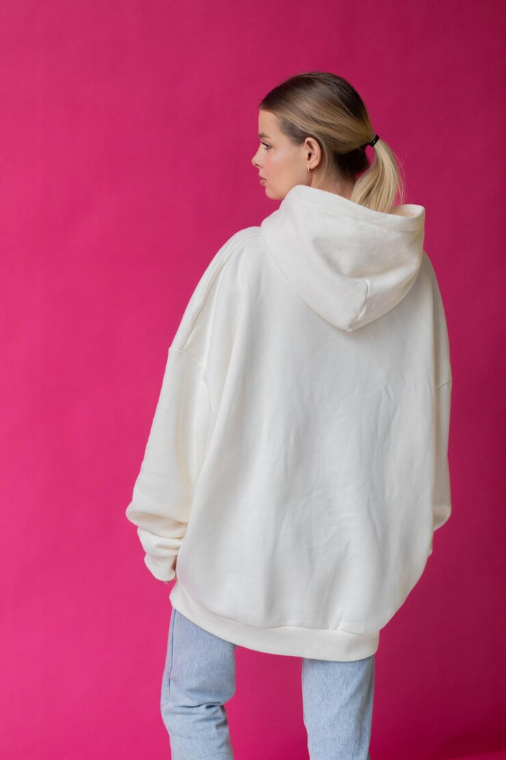 Mooie blonde vrouw in een witte hoodie en blauwe spijkerbroek die op een roze achtergrond poseert
