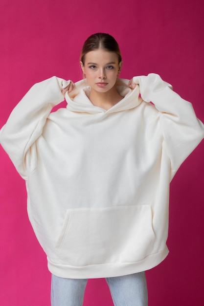 Mooie blonde vrouw in een witte hoodie en blauwe spijkerbroek die op een roze achtergrond poseert