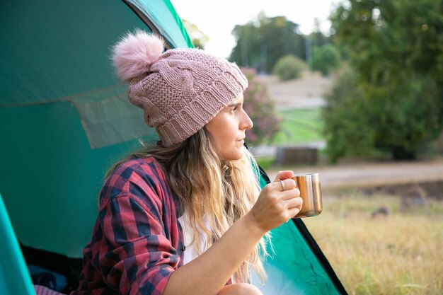 Mooie blonde vrouw in hoed die thee drinkt, in tent zit en op landschap kijkt. Kaukasische langharige reiziger die kop houdt of in park ontspant. Toerisme, reis en vakantieconcept