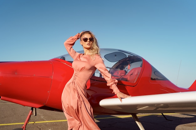Mooie blonde vrouw met golvend haar, gekleed in een stijlvolle roze jurk van zijde, poserend in de buurt van een privévliegtuig