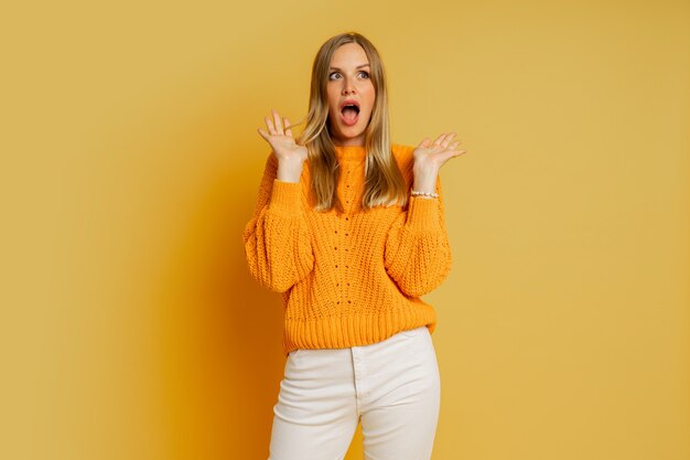 Mooie blonde vrouw met verrassingsgezicht in oranje stijlvolle herfsttrui die zich voordeed op geel.