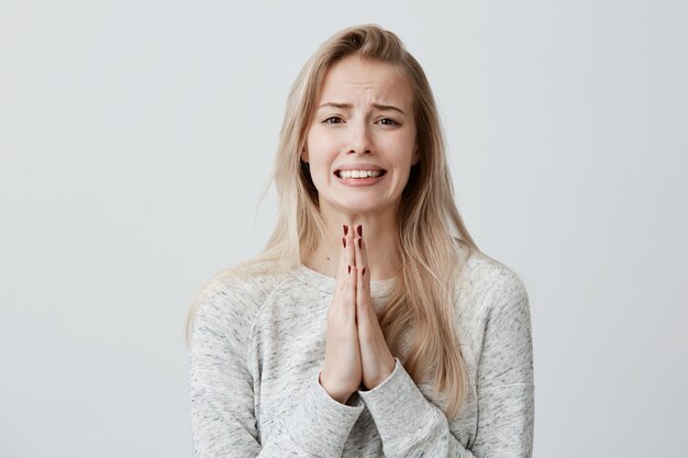 Mooie blonde vrouwelijke balde tanden met bedelende blik met handpalmen tegen elkaar gedrukt, vragend om medelijden