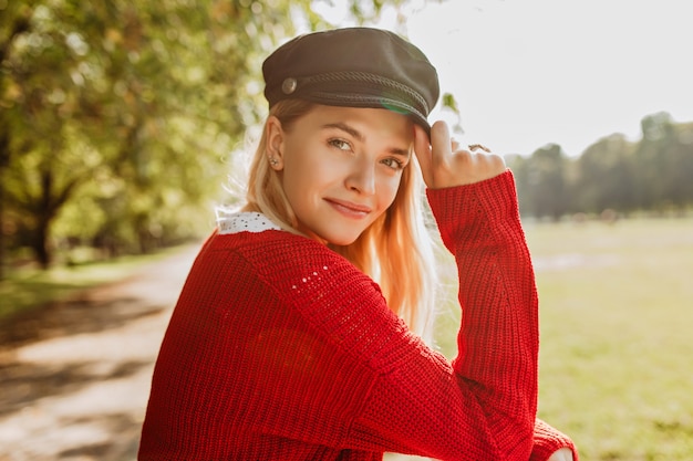 Mooie blonde ziet er mooi uit in rode trui en trendy zwarte hoed in het park. Glimlachend meisje geniet van zonnige herfstdag.