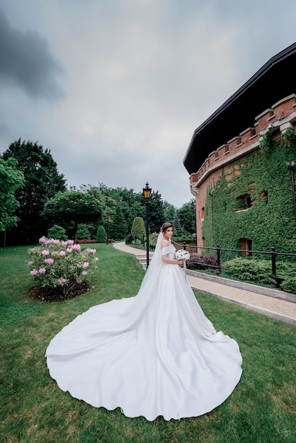Mooie bruid met bruiloft boeket en luxe trouwjurk staat voor een gebouw bedekt met groene bladeren