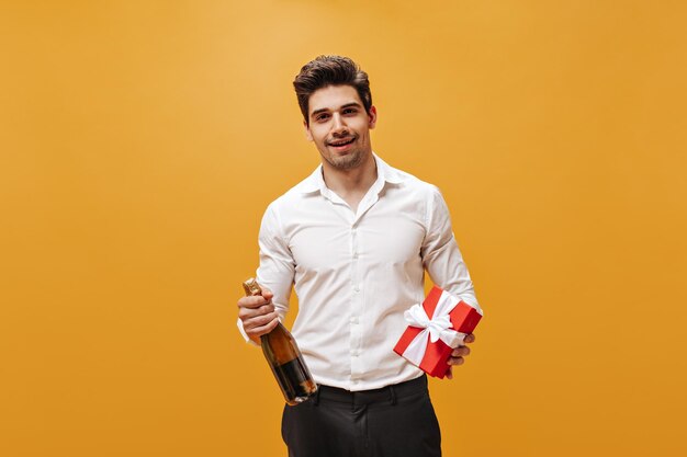 Mooie brunet man in wit stijlvol shirt en zwarte broek glimlacht houdt champagnefles en rode geschenkdoos op oranje achtergrond