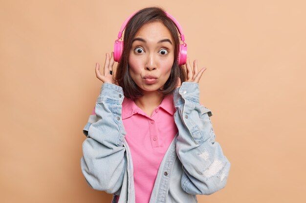 Mooie brunette Aziatische vrouw met donker haar houdt lippen gevouwen heeft geschokte uitdrukking luistert muziek via roze draadloze koptelefoon draagt spijkerjasje geïsoleerd over beige muur