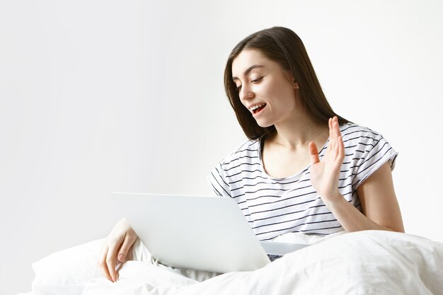 mooie brunette blanke vrouw van in de twintig zittend op wit beddengoed in de slaapkamer en videoconferenties