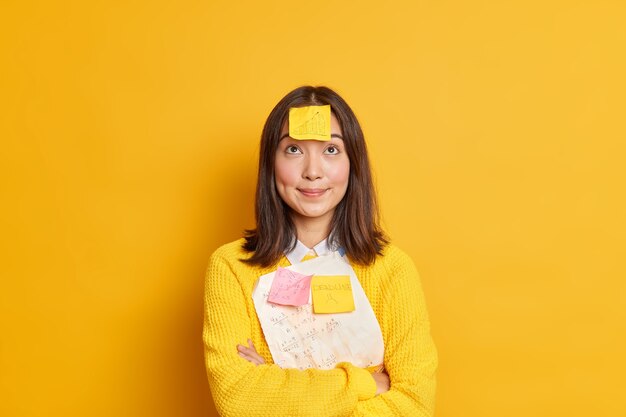 Mooie brunette jonge Aziatische studente probeert materiaal te onthouden voor examen heeft deadline memosticker geplakt op voorhoofd geconcentreerd boven staat gekruiste handen