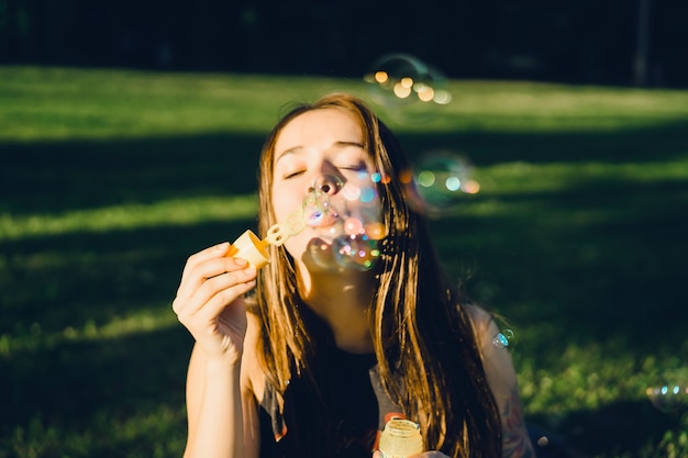 Gratis foto mooie brunette meisje met lang haar puffs zeepbellen in het park