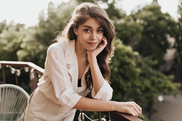 Mooie brunette vrouw in beige outfit poseren op terras Aantrekkelijk krullend meisje in blouse op zoek naar camera op balkon