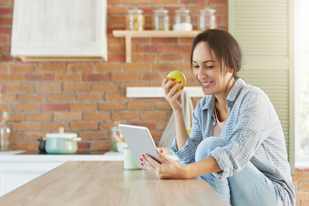 Mooie brunette vrouw in casual kleding, zit in de keuken, eet appel, maakt gebruik van moderne tablet,