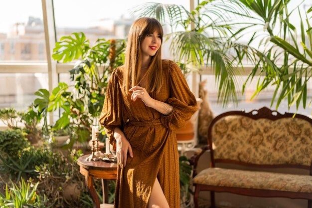 Mooie brunette vrouw in elegante jurk die geniet van de ochtend in een stijlvolle Boheemse woonkamer met huisplanten