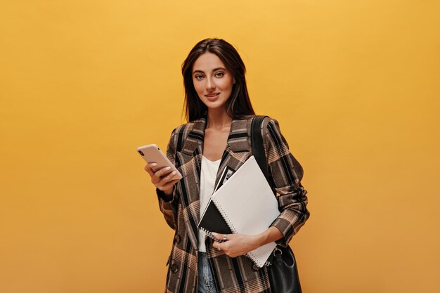 Mooie brunette vrouw in wit T-shirt en stijlvol jasje houdt telefoon en notebooks Aantrekkelijk meisje in modieuze outfit kijkt naar de camera op gele achtergrond