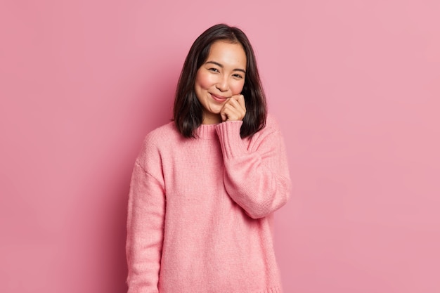 Mooie brunette vrouw met oosterse uitstraling houdt hand op gezicht heeft zachte glimlach kijkt tevreden naar camera heeft kuiltjes op rouge wangen poses