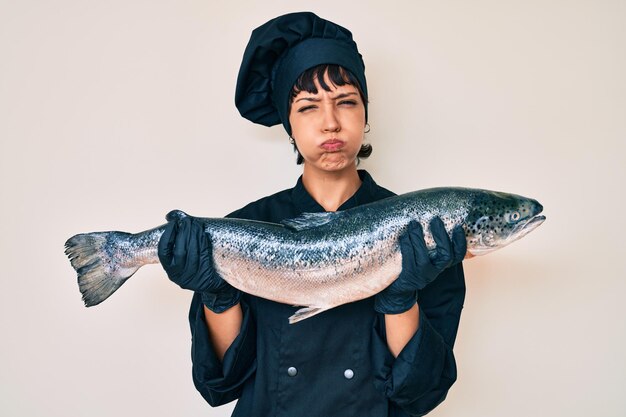 Mooie brunette vrouw professionele chef-kok met verse zalm vissen puffende wangen met een grappig gezicht. mond opgeblazen met lucht, lucht vangen.