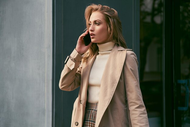 Mooie casual blond meisje in trenchcoat praten op mobiele telefoon bedachtzaam wegkijkend buiten