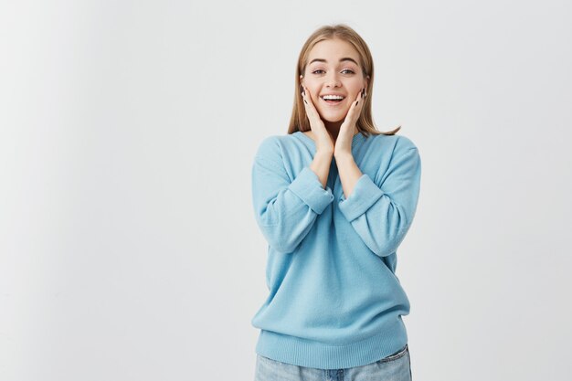 Mooie charmante blonde blanke meisje draagt blauwe trui en spijkerbroek wijd open mond, zegt Wow, opgewonden verbaasd blik, hand in hand op haar gezicht, blij met onverwacht cadeau