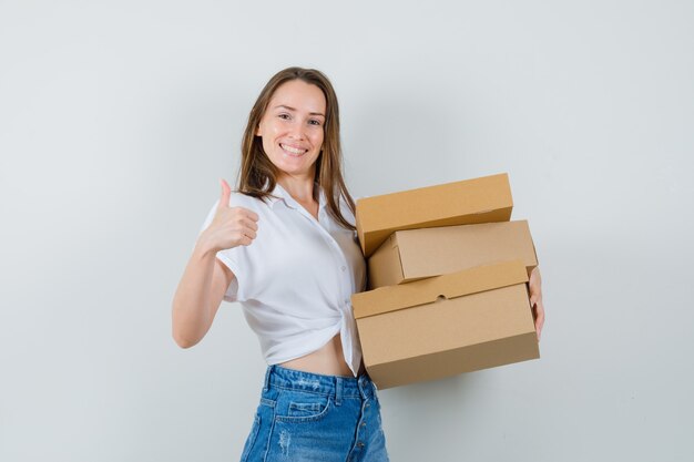 Mooie dame die dozen houdt terwijl duim in witte blouse wordt getoond en tevreden, vooraanzicht kijkt.