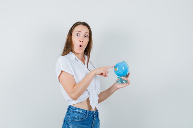 Mooie dame die ergens op wereldbol in witte blouse wijst en verbaasd kijkt. vooraanzicht.