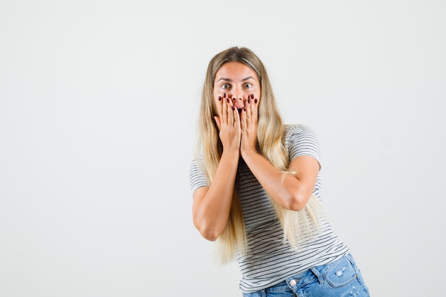 Mooie dame die in t-shirt hand op haar wangen houdt en bang, vooraanzicht kijkt.