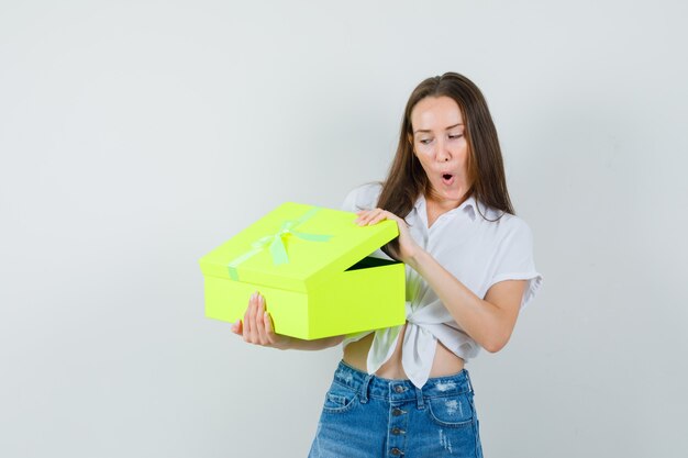 Mooie dame geschenkdoos openen in witte blouse, spijkerbroek en op zoek verrast, vooraanzicht.