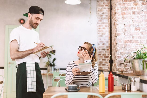 Mooie dame in bril zittend aan tafel met menu in handen en bedachtzaam kijkend naar ober tijdens het bestellen in café