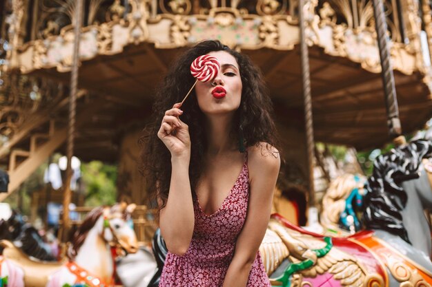 Mooie dame met donker krullend haar in jurk die staat en haar oog bedekt met lolly pop snoep terwijl ze dromerig in de camera kijkt met carrousel op de achtergrond