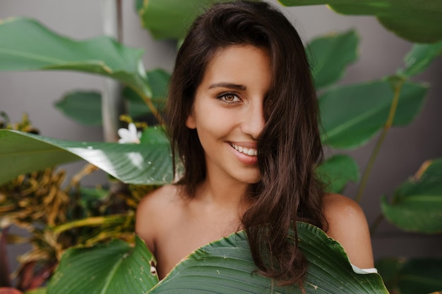 Mooie dame met krullend haar en groene ogen lacht lief Vrouw met lange wimpers poseren tegen de achtergrond van tropische plant