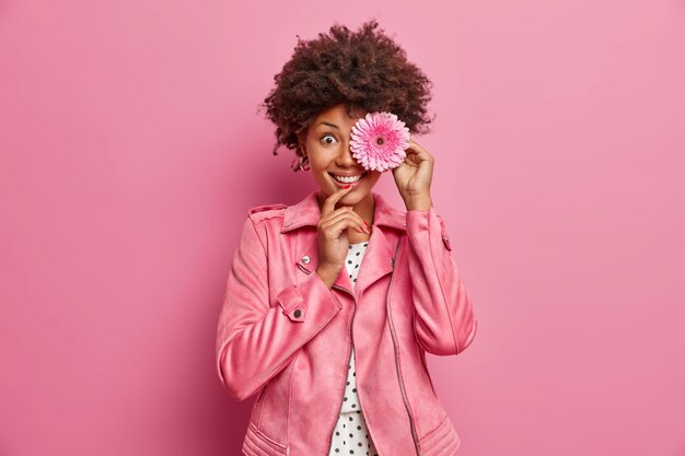 Mooie dame met krullend haar met brede glimlach houdt roze gerberabloem voor de ogen, plukt bloemen van bloeiende lenteweiden, gekleed in roze jasje, gaat krans maken