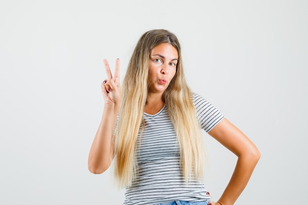 Mooie dame met v-sign terwijl ze haar lippen in t-shirt pruilt en er klaar voor kijkt. vooraanzicht.