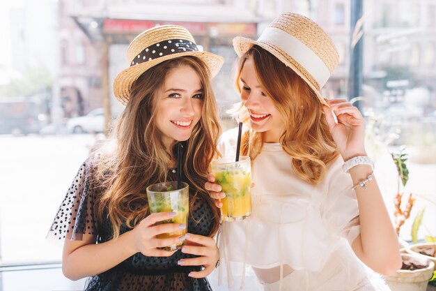 Mooie dames dragen soortgelijke strohoeden die samen plezier hebben en genieten van ijskoude fruitcocktails in de zomerdag