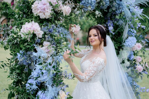 Mooie donkerbruine bruid dichtbij de overwelfde galerij die van blauwe hortensia en ruscus wordt gemaakt, huwelijksdag