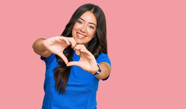 Mooie donkerbruine jonge vrouw die vrijetijdskleding draagt die in liefde glimlacht die de vorm van het hartsymbool met handen doet. romantisch begrip.