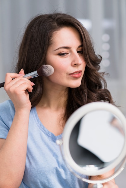 Mooie donkerbruine vrouw met borstel en spiegel die make-up doet