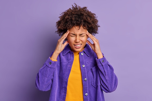 Mooie donkere huid Afro-Amerikaanse vrouw raakt tempels heeft ondraaglijke hoofdpijn lijdt aan migraine gekleed in modieus fluwelen jasje.