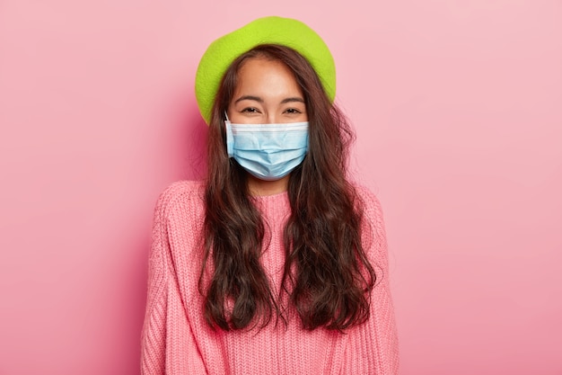 Mooie donkerharige Aziatische dame heeft een epidemische ziekte, draagt een beschermend medisch masker, een groene baret en een trui