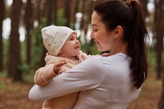 Mooie donkerharige vrouw met witte kleding die buiten poseert, baby baby in handen houdt en met grote liefde naar dochter kijkt, spelend in het bos